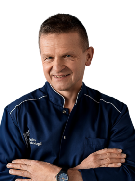 Portret dr. Jerzego Leszczyńskiego, chirurga naczyniowego z Kliniki Flebologii. Lekarz o krótkich, ciemnych włosach uśmiecha się łagodnie, mając skrzyżowane ramiona. Jest ubrany w granatowy uniform medyczny z białym logo kliniki na piersi. Zdjęcie na czystym, białym tle.