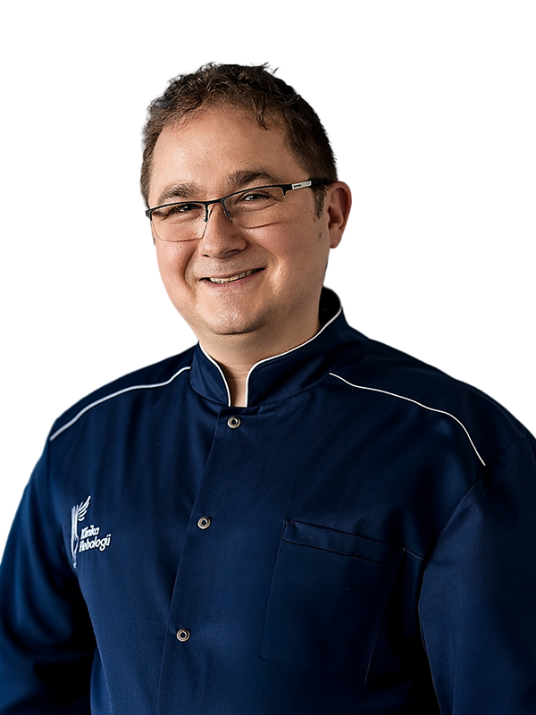 Portret doktora Krzysztofa Celejewskiego, chirurga i flebologa z Kliniki Flebologii. Lekarz o krótkich, ciemnych włosach uśmiecha się łagodnie, mając skrzyżowane ramiona. Jest ubrany w granatowy uniform medyczny z białym logo kliniki na piersi. Zdjęcie na czystym, białym tle.