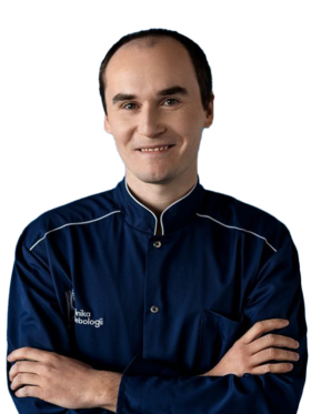 Portret Piotra Błachowicza, chirurga i flebologa z Kliniki Flebologii w Warszawie. Mężczyzna o krótkich ciemnych włosach uśmiecha się do aparatu, mając skrzyżowane ramiona. Ubrany jest w granatowy kitel medyczny z białymi lamówkami oraz widocznym logo „Klinika Flebologii” na piersi. Tło jest jednolite i białe.