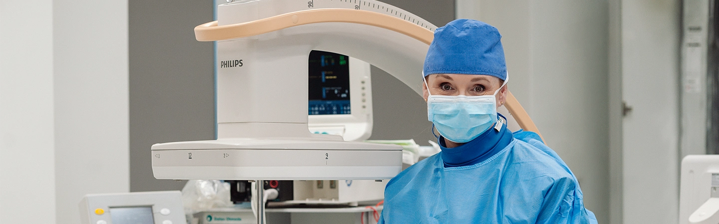 Lekarz anestezjolog w sterylnej odzieży ochronnej i masce, przygotowujący się do zabiegu w nowoczesnej sali operacyjnej wyposażonej w ramię C Philips do precyzyjnej diagnostyki naczyniowej.